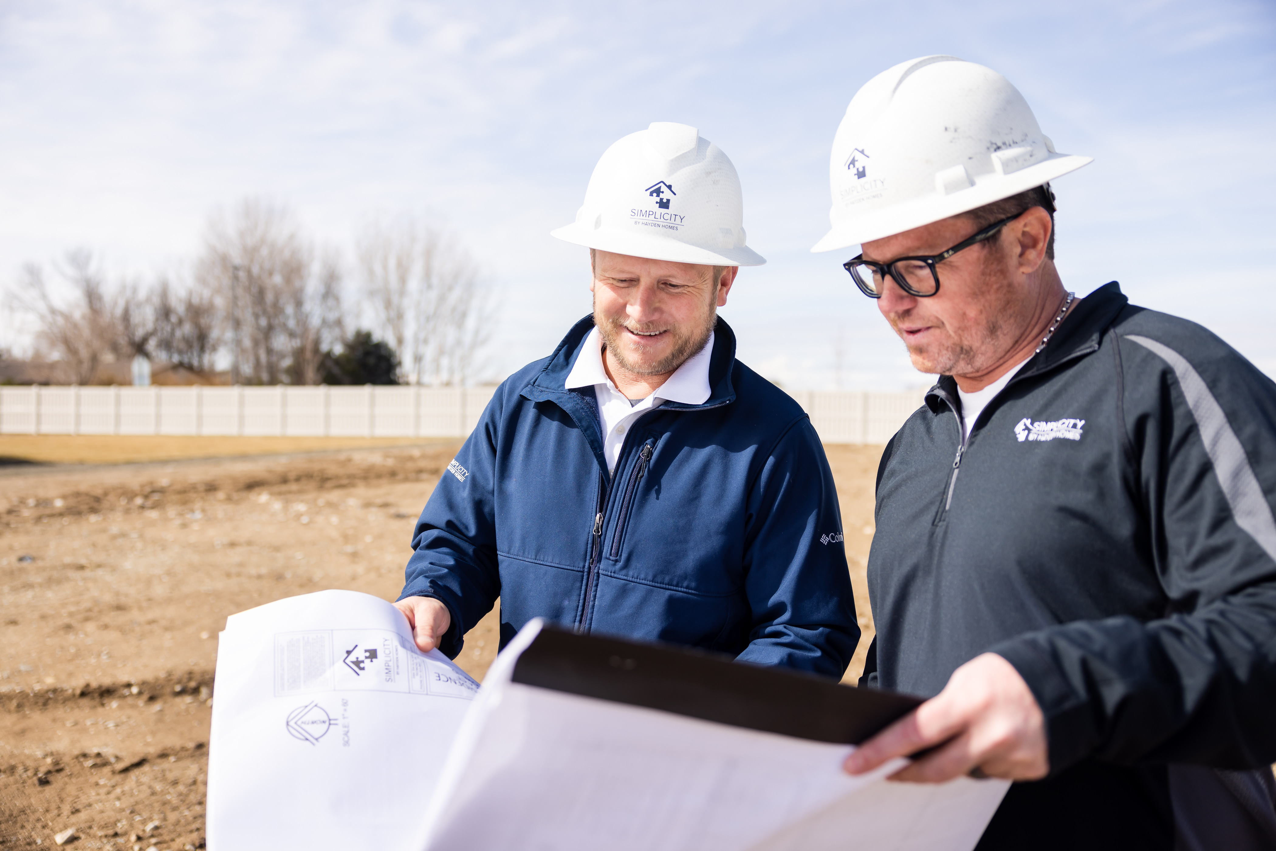 How to Buy Land to Build a New Home. Close up on two Simplicity Homes employees in hard hats reviewing building plans